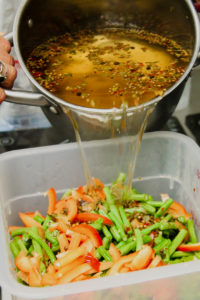 Action photo of a light brown pickling broth being poured into a tub of bright red peppers and green beans.