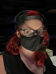 Photo of a woman in a black face mask, backwards baseball cap, and headset. She wears thick rim glasses and has curly red hair. She looks at the camera while a downwards light lights up her face.