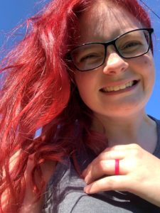 Selfie of a woman smiling with bright red hair. She wears a grey shirt, thick rim glasses, and a hot pink wedding ring.
