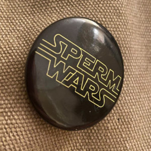 Photo of a black button with yellow text that reads "SPERM WARS" in font similar to the Star Wars logo. The button is on a cream fabric background.