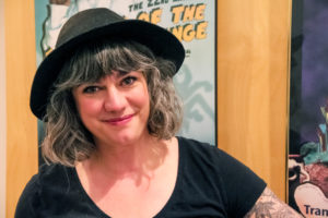 Phot of Megan Dart, a white woman with curly hair, wearing a black hat and shirt. She smiles at the camera with Fringe posters in the background.