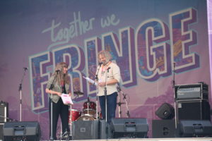 Megan and Murray smiling at each other on the ATB Main Stage in August 2021, with the large Together We Fringe design in the background.