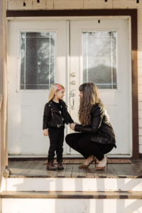 Photo of Megan Dart in all black with a leather jacket, on a porch outside of a building with two white doors. She kneels beside her daughter Alice, who is also wearing all black with a leather jacket, as they look at each other. Photo by Mat Simpson