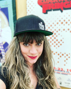 Photo of Megan Dart, with long hair, smiling at the camera with a black Fringe Theatre hat. Fringe posters line the background.
