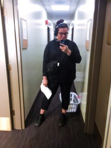 Mirror selfie of Lindy Mullen, who wears all black with her dark hair tied back in a messy bun. In her left hand is her phone and in her right is a script. She wears a satchel and headset.