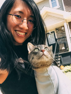 Yu-Chen, a woman with long black hair and glasses. She smiles at the camera and her grey cat looks up at her.