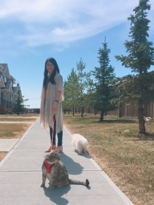 Yu-Chen, a woman with long black hair and glasses. She wears jeans and a long cardigan while walking her two pets on a sunny day.