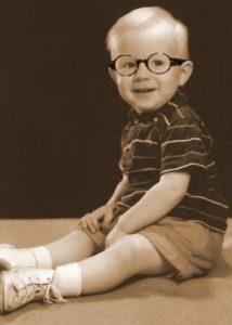 Sepia toned photo of Gerald Osborn as a young child, wearing a stripped t-shirt, shorts, long white socks and white shoes with thick rimmed black glasses. His platinum blonde hair is styled with a side part.