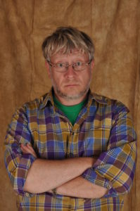 Gerald Osborn, his arms crossed in front of him with sort of messy hair and glasses. He wears a multi-coloured plaid shirt and green t-shirt underneath.