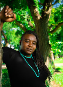 Shima Robinson leans against a tree wearing a black turtleneck and a jade necklace.
