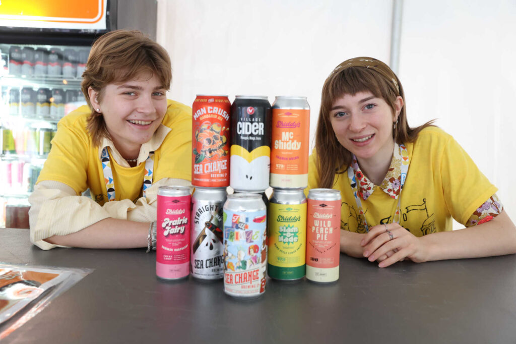 Two volunteers posing at a countertop in front of a stack of various Sea Change beverages