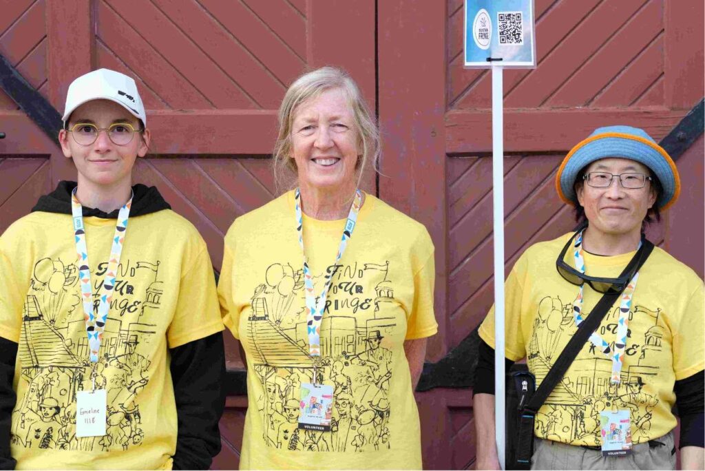 Three front of house Volunteers standing outside a venue holding a sign on a long stick