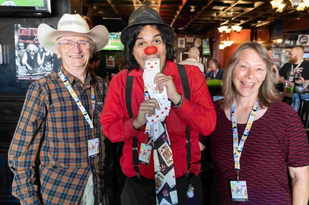 Two Fringe billet hosts standing next to a clown as the clown pretends to vomit a deck of cards out their mouth.