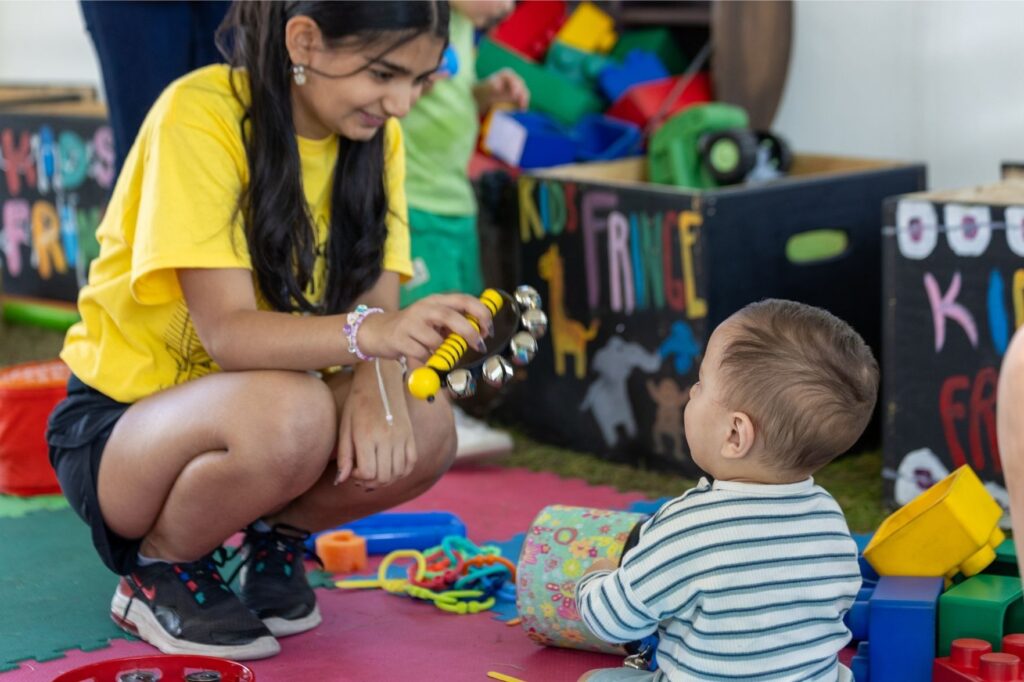 A volunteer crouching in front of a young child shaking a toy with bells on it