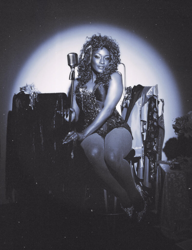 A woman with long curly hair sits on a chair behind an old-fashioned stand microphone.