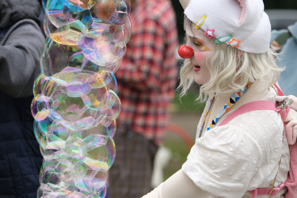 A clown with a white shirt, pink backpack and a bunny ear toque plays with bubbles.