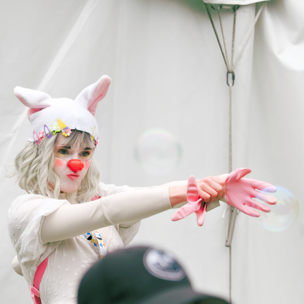 A clown dressed in white, wearing a floppy eared toque pulls on a pair of pink gloves.