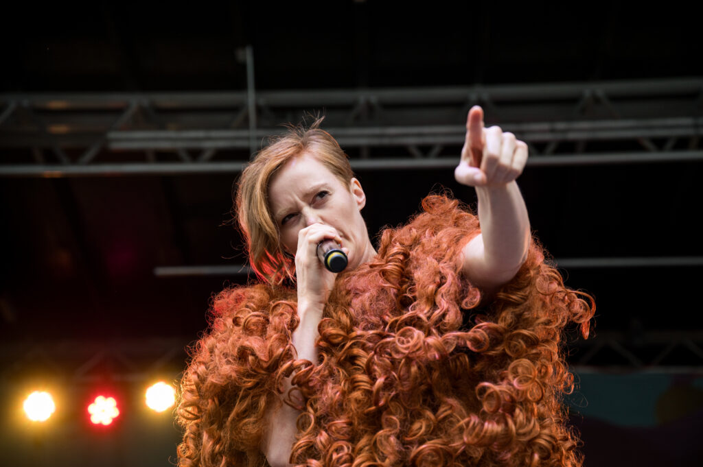 A performer wearing a giant wig speaks into a microphone and points towards the audience.