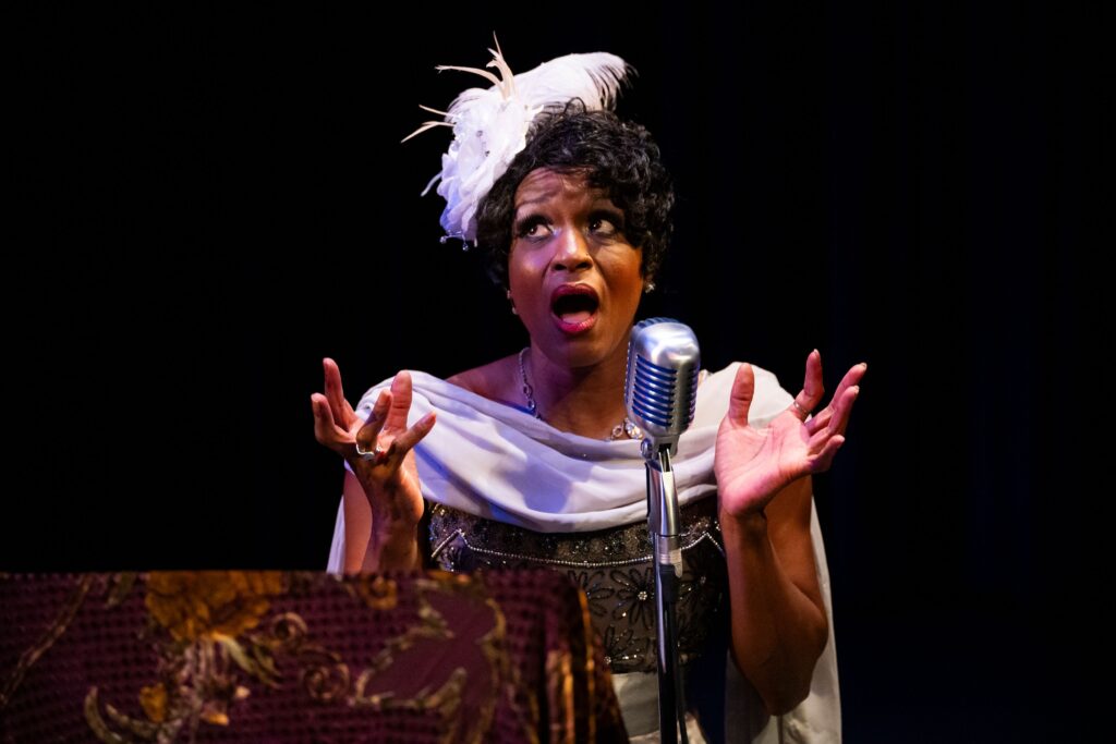 A woman in a feathered hat sings into an old-fashioned stand microphone.