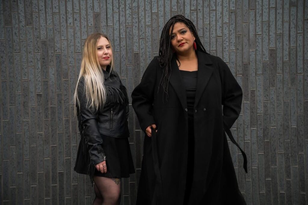 Two artists wearing leather and black pose against a dark background.