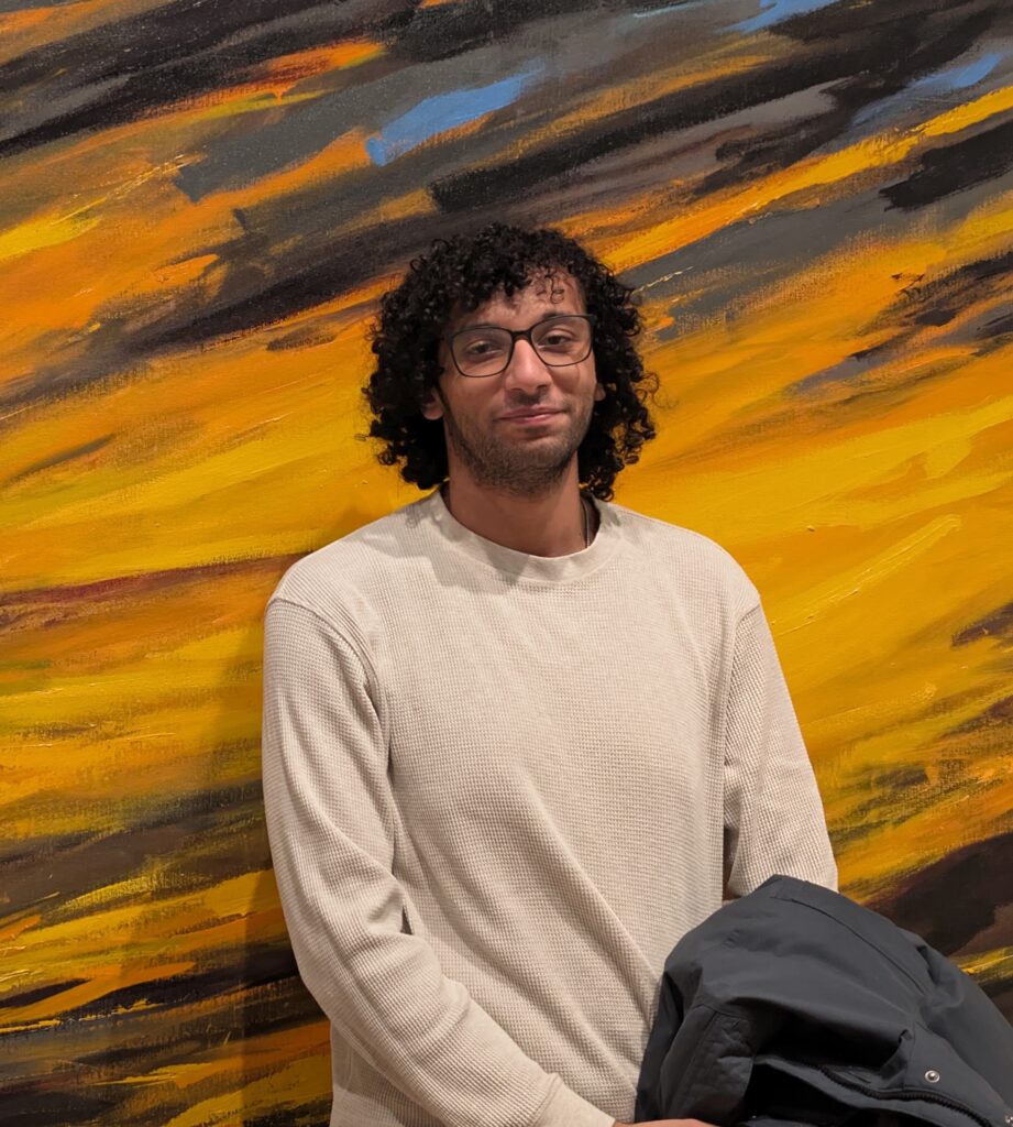 A man with curly hair and glasses stands in front of an orange, yellow, black, and blue-painted wall.
