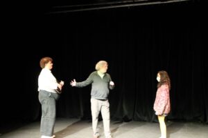 Director, Murray Utas stands onstage with the actors. He has his arm stretched out as he talks to them.