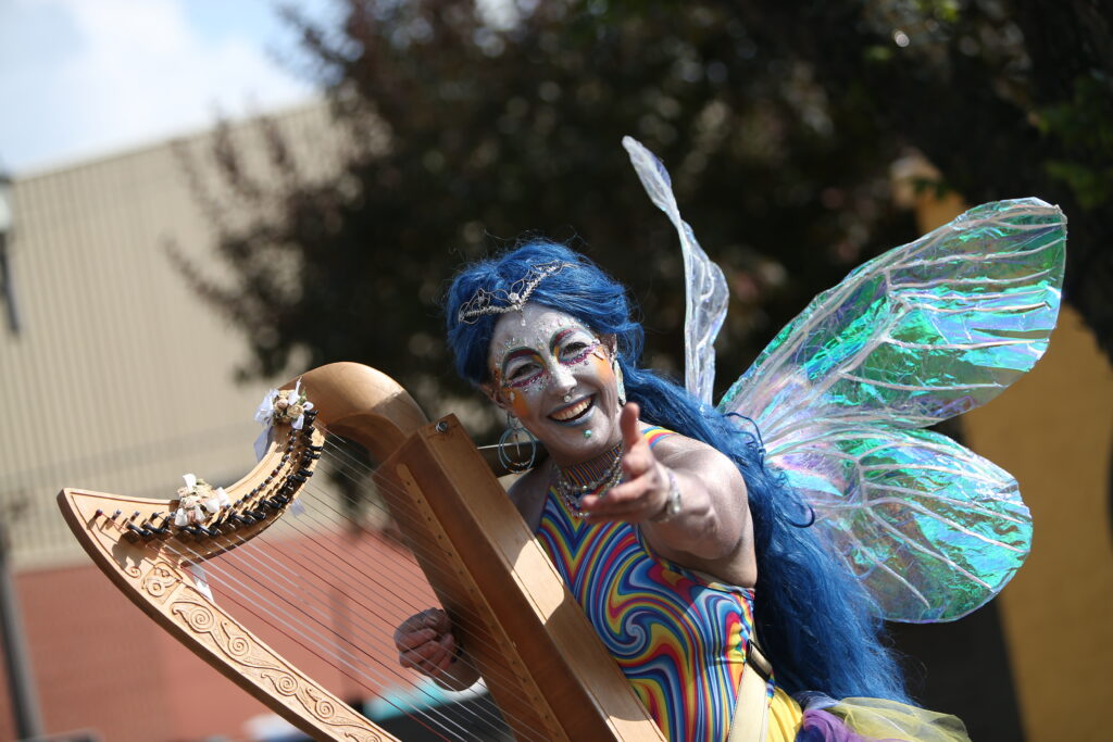 A busker dressed as a fairy holds a harp and reaches her hand out to the camera.