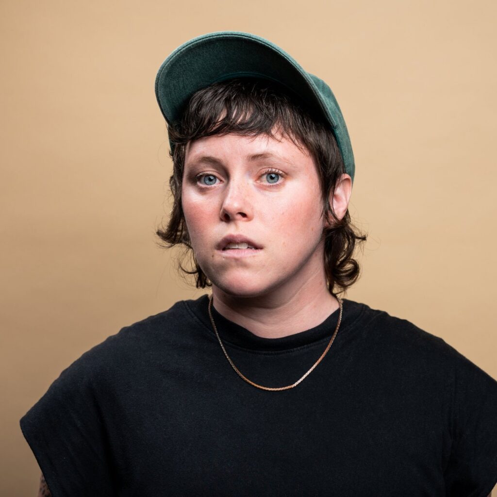 A headshot of Elena Belyea wearing a green ballcap, black shirt, and gold chain necklace.