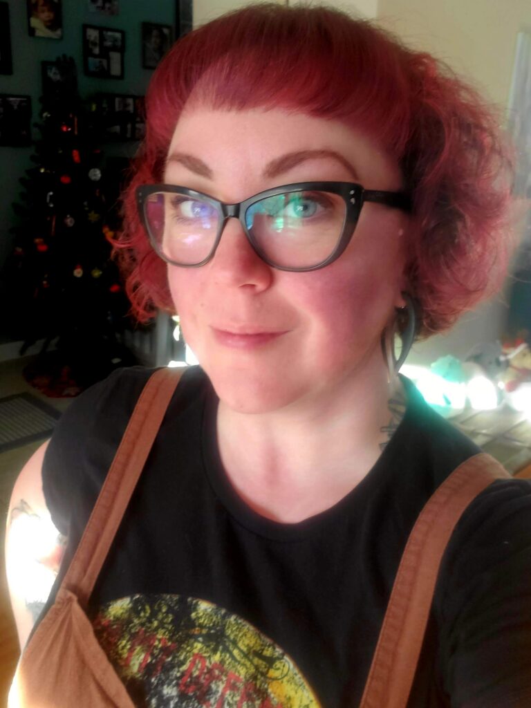 A headshot of Kat Evans wearing large glasses, a black shirt, and brown overalls.