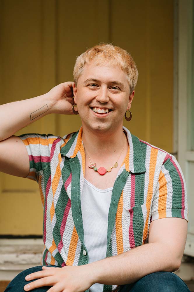 A headshot of Mac Brock wearing a green, yellow, and while vertically-striped shirt.