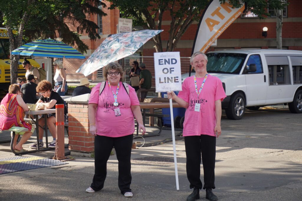 Two Fringe volunteers in pink shirts and one holding a tall sign that says 'End Of Line'