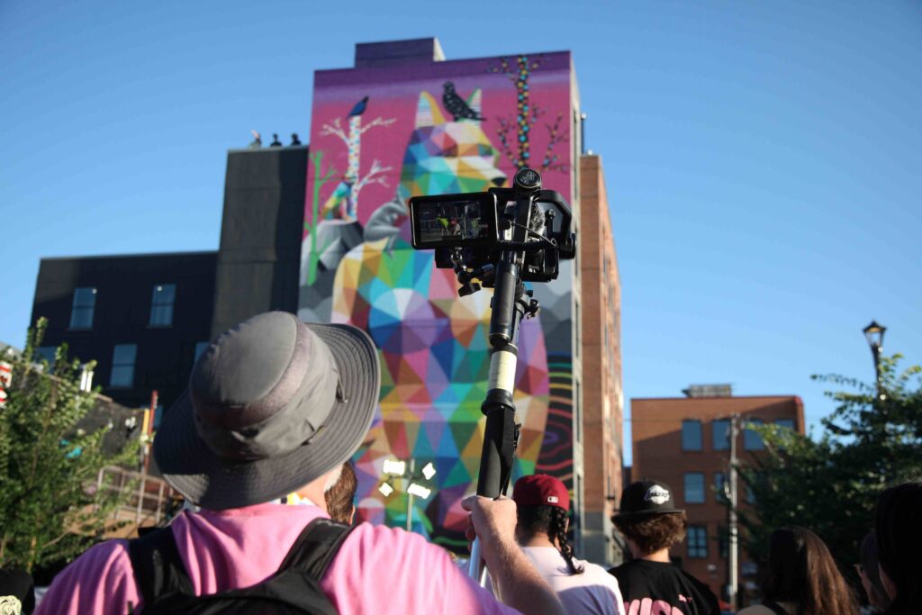 A Fringe Multimedia volunteer in a pink shirt from the shoulders up holding up a camera on a Tripod
