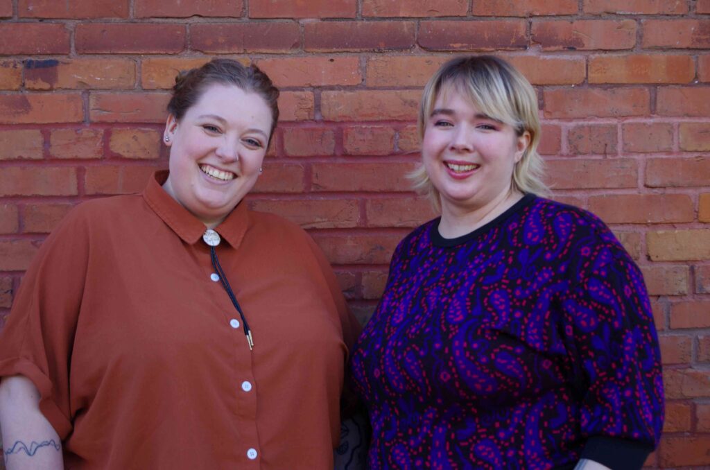 Two Fringe Volunteer Department staff members (Olivia Bogaard and Rain Bossert) smiling at the camera for a photo in front of a red brick wall