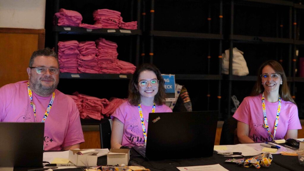 Three Fringe VHQ Volunteers in pink Fringe shirts working at computers behind a desk and smiling for a photo