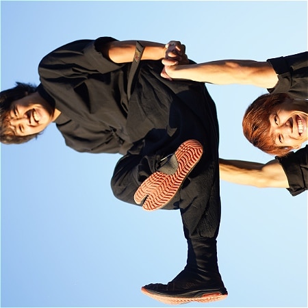 Side-ways image. Two performers. One balances on the others hands.