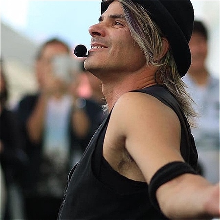 A performer in all back with long hair and a microphone poses for the camera.